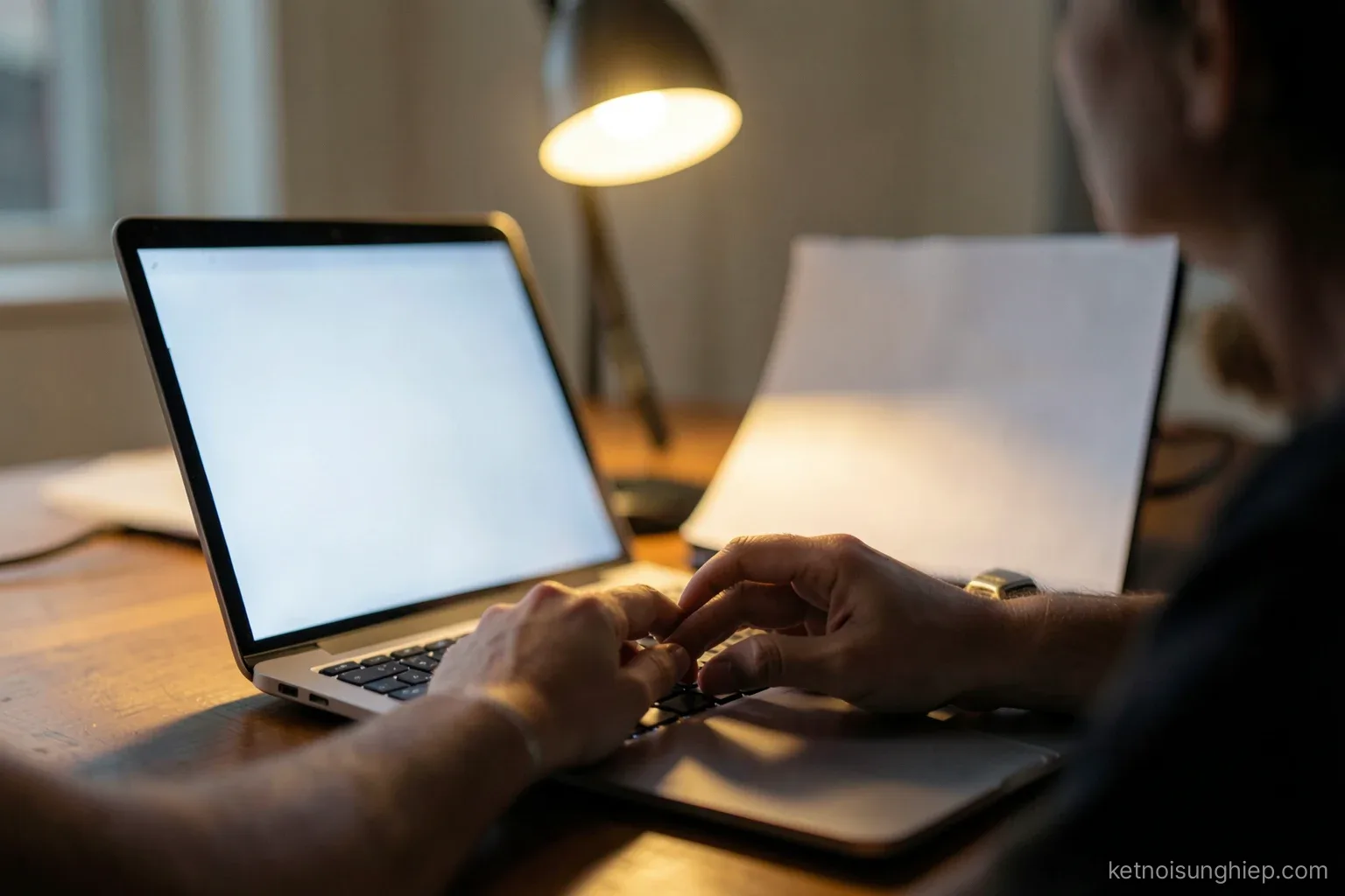 Close-up of hands typing on a laptop with a blank screen at a quiet office desk in early evening, desk lamp glowing