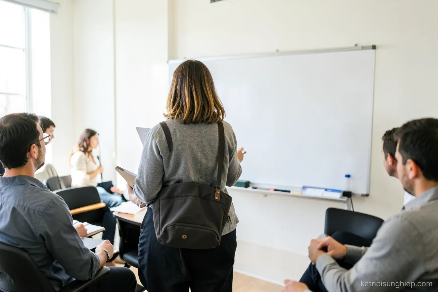 A woman presents to a small team in a bright meeting room, standing beside a blank whiteboard, seen from the back of the room