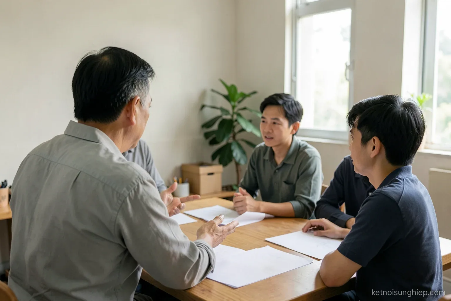 A Vietnamese man in his late forties seen from behind, explaining something to two younger colleagues around a workshop table with blank papers, soft daylight