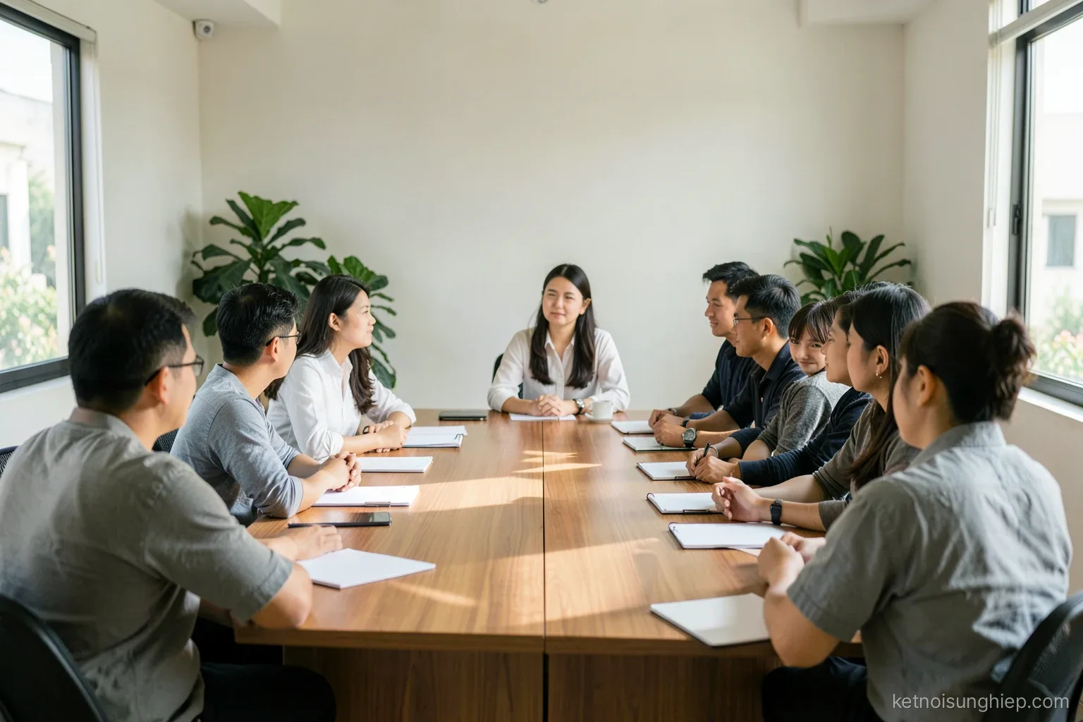 Wide shot of a mixed-age Vietnamese team around a long meeting table in a sunlit office, people in profile and from behind, blank notebooks